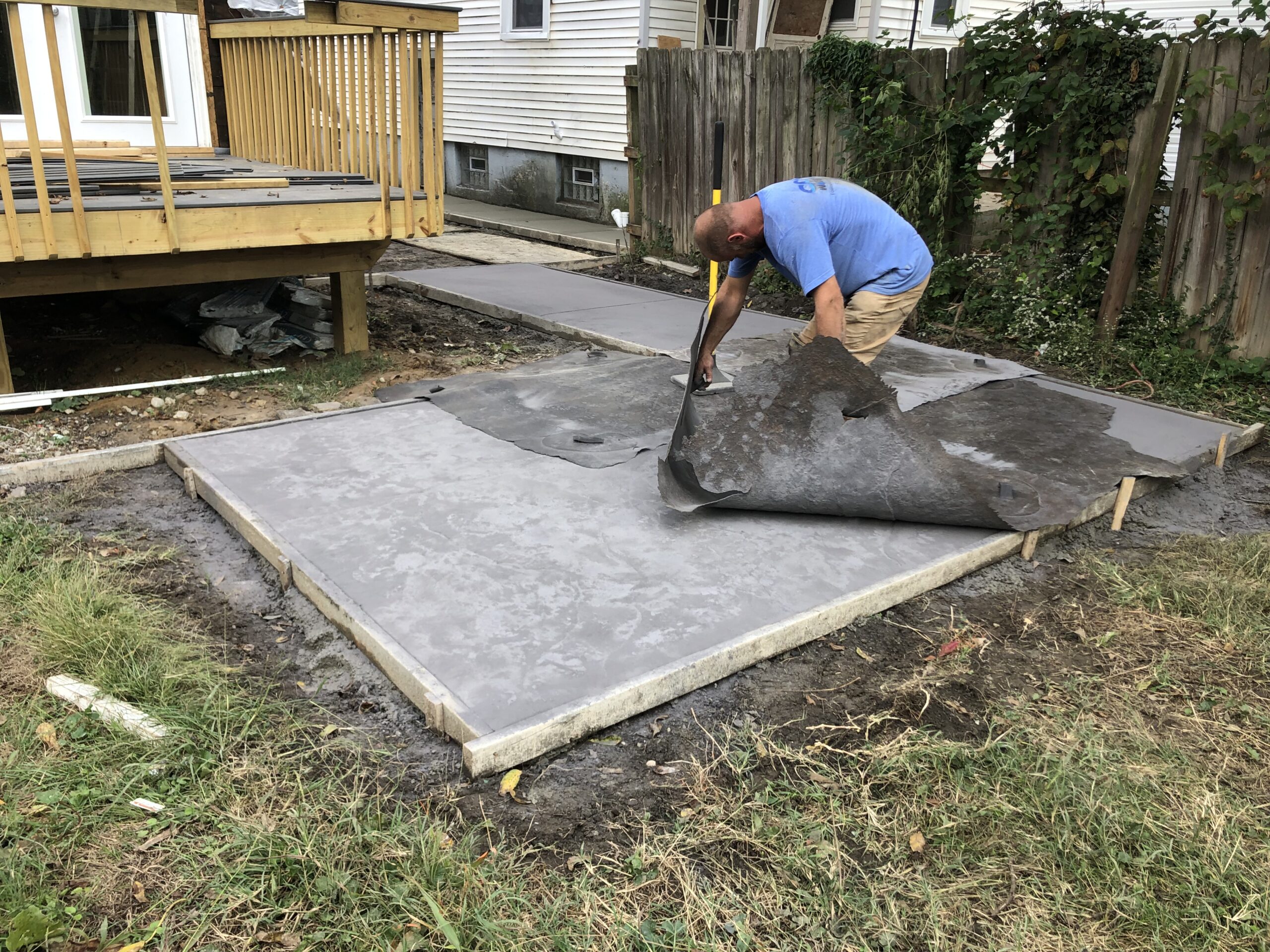 A man removes a sheet of plastic from a freshly poured concrete slab in a backyard, with a wooden deck and fence in the background.