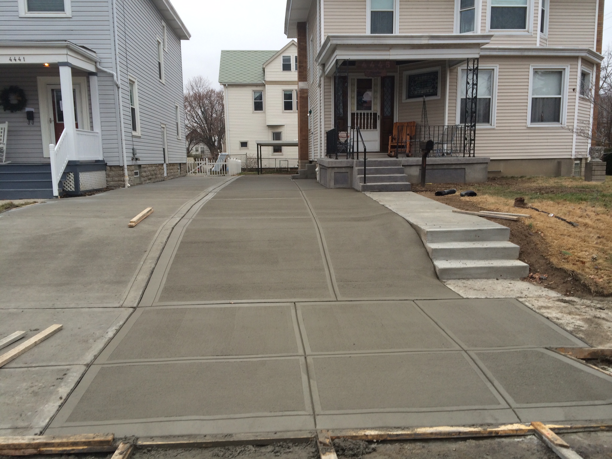 Freshly poured concrete driveway and steps in front of a residential house, bordered by adjacent houses and a patch of grass.