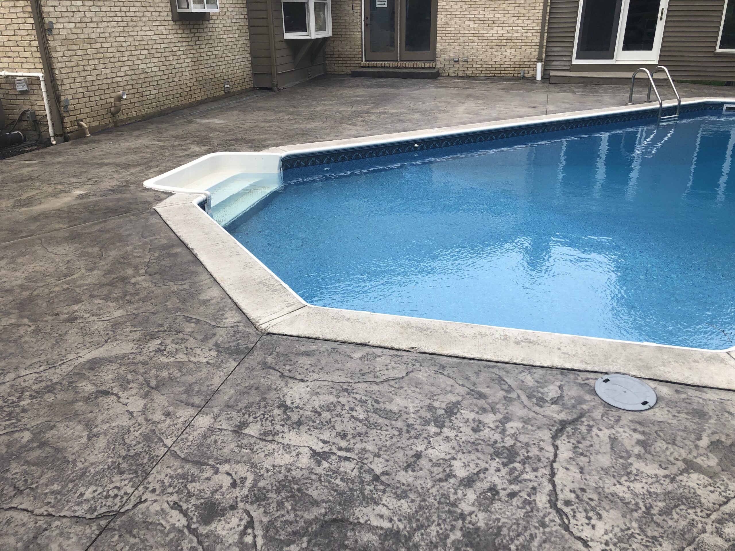 A backyard swimming pool with a concrete patio, metal ladder, and shallow steps leading into the water near a beige brick house.