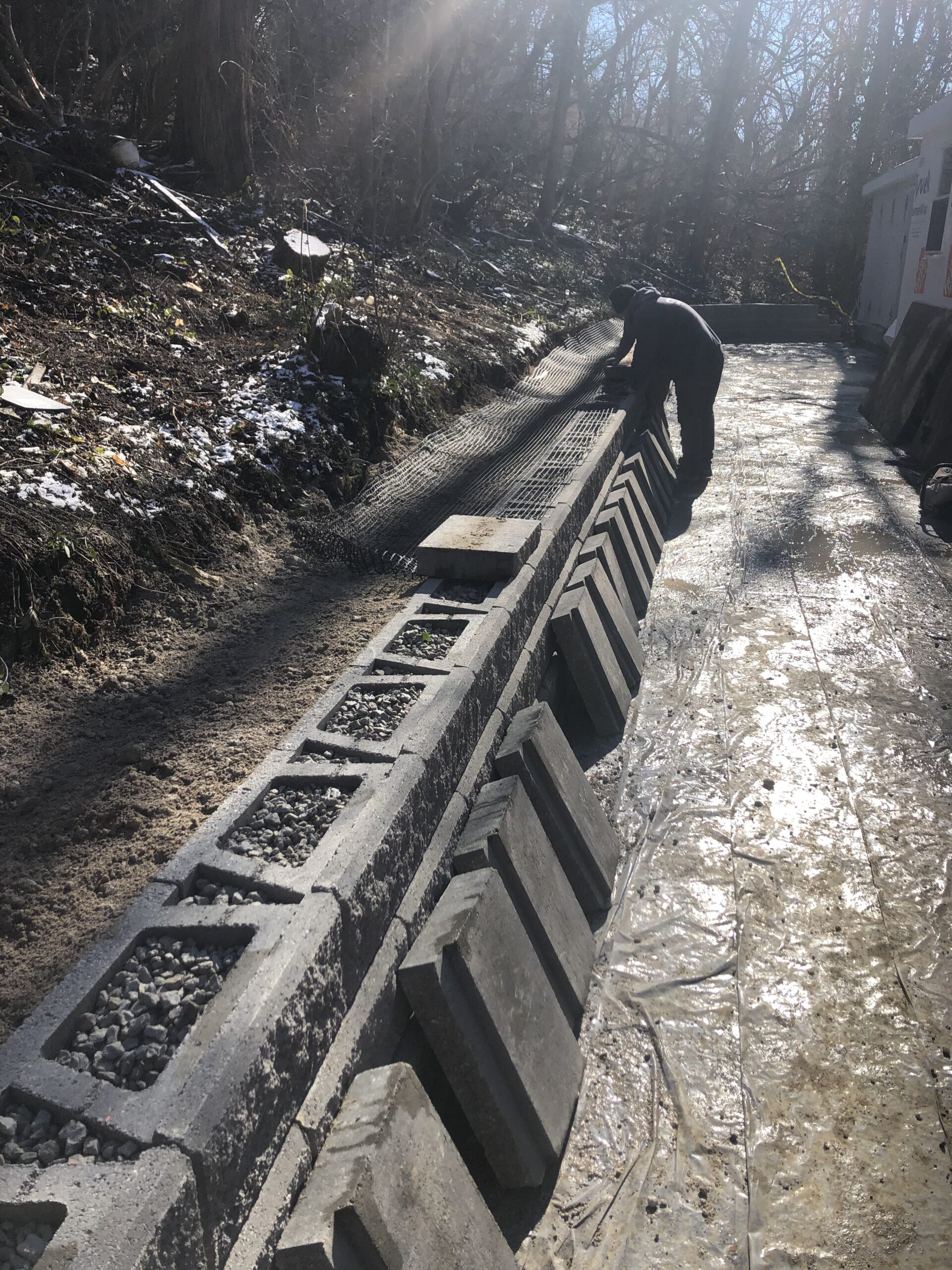 A person installs concrete blocks to construct a retaining wall along a sloped, partially wooded area on a sunny day.
