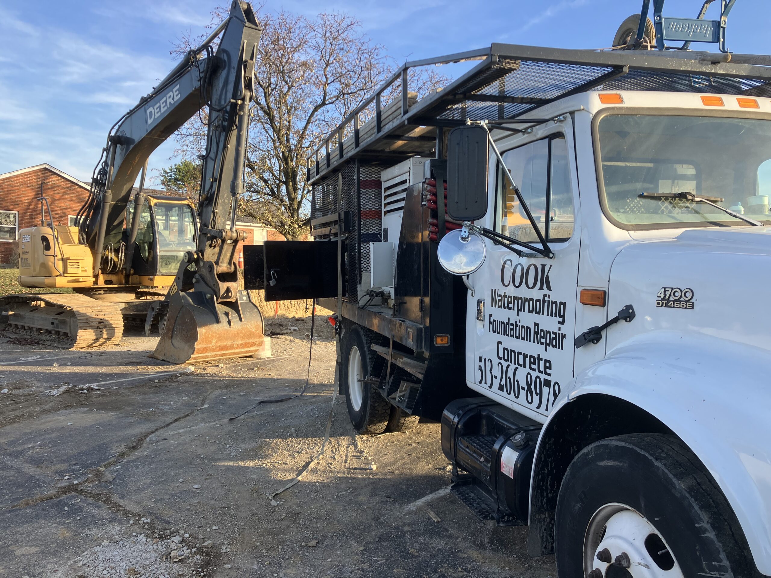 A white Cook Waterproofing service truck with equipment is parked next to a large excavator at a construction site on a sunny day.