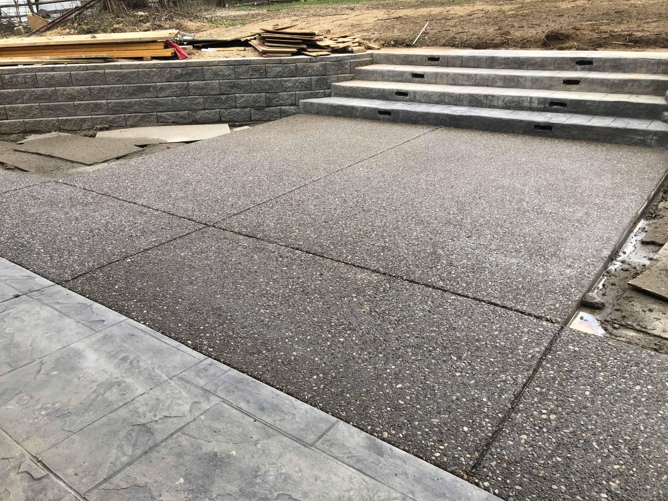A recently poured exposed aggregate concrete patio with steps and retaining walls, surrounded by some unfinished landscaping and construction materials.