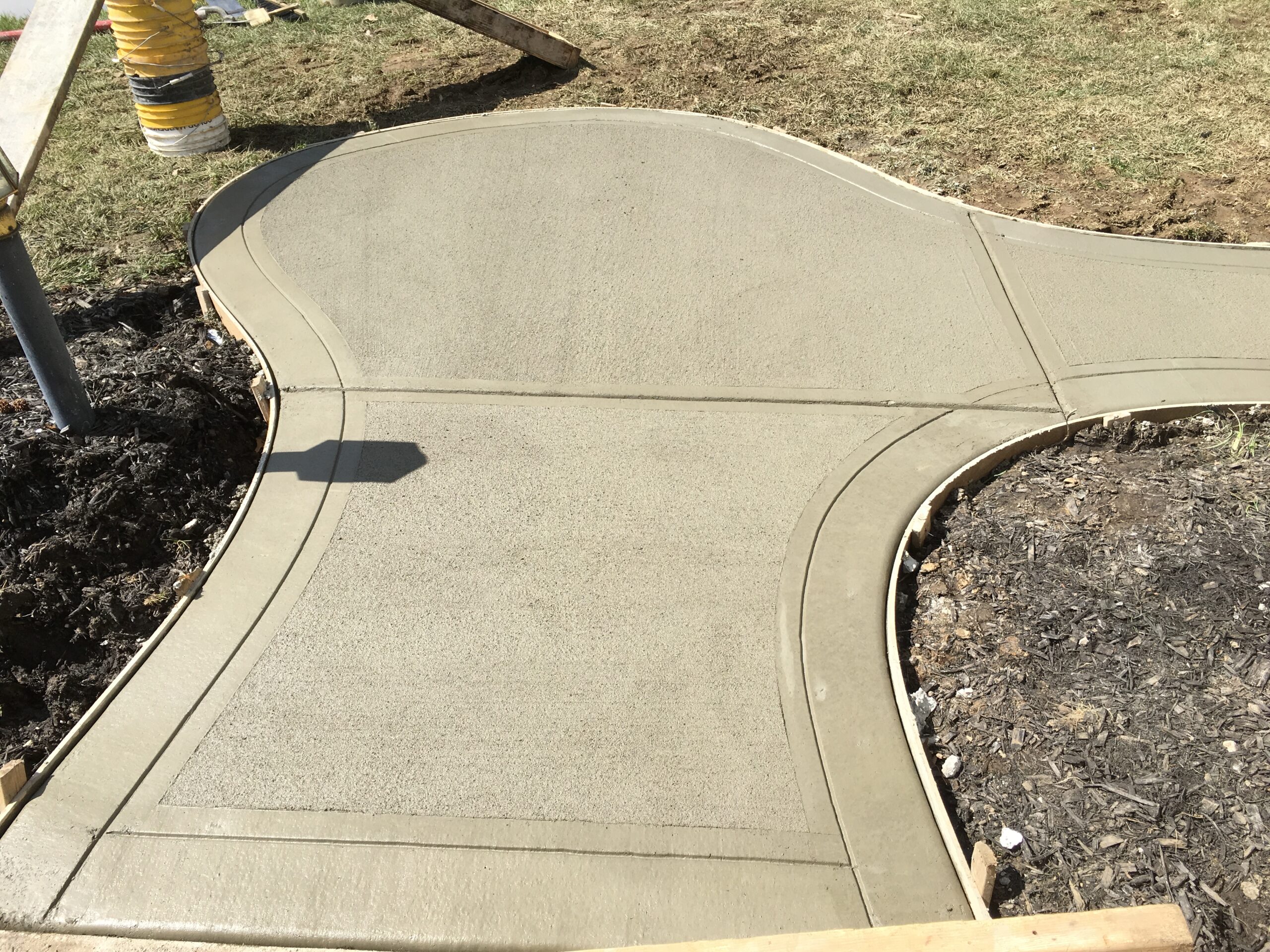 Freshly poured concrete sidewalk in a curving shape, bordered by soil and mulch, with construction materials visible on the side.
