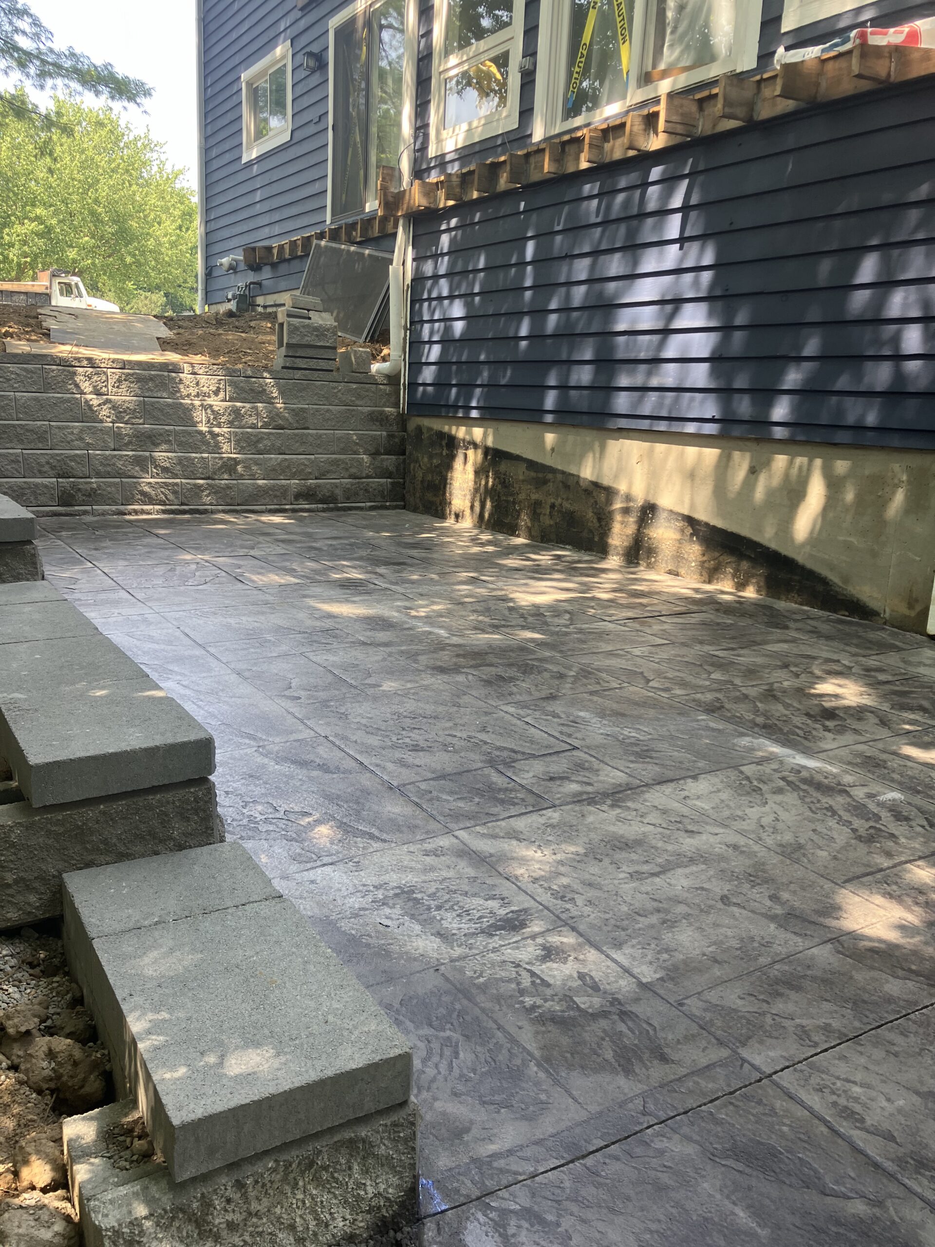 A newly constructed stone patio with stairs leads up to a house with blue siding. The patio and steps appear freshly built, with construction still in progress.
